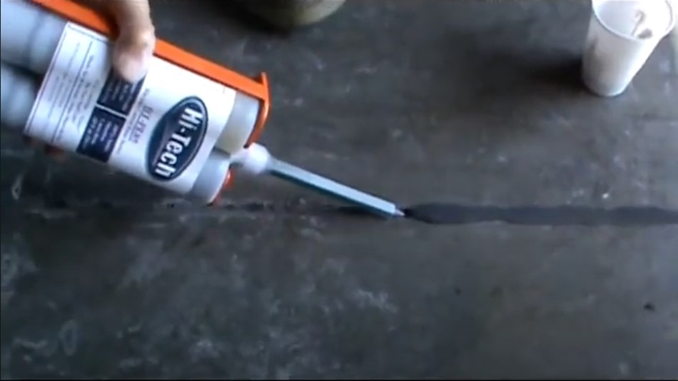 A person's hand holds a caulking gun dispensing a dark sealant into a crack on a concrete surface. A small plastic cup is visible nearby.