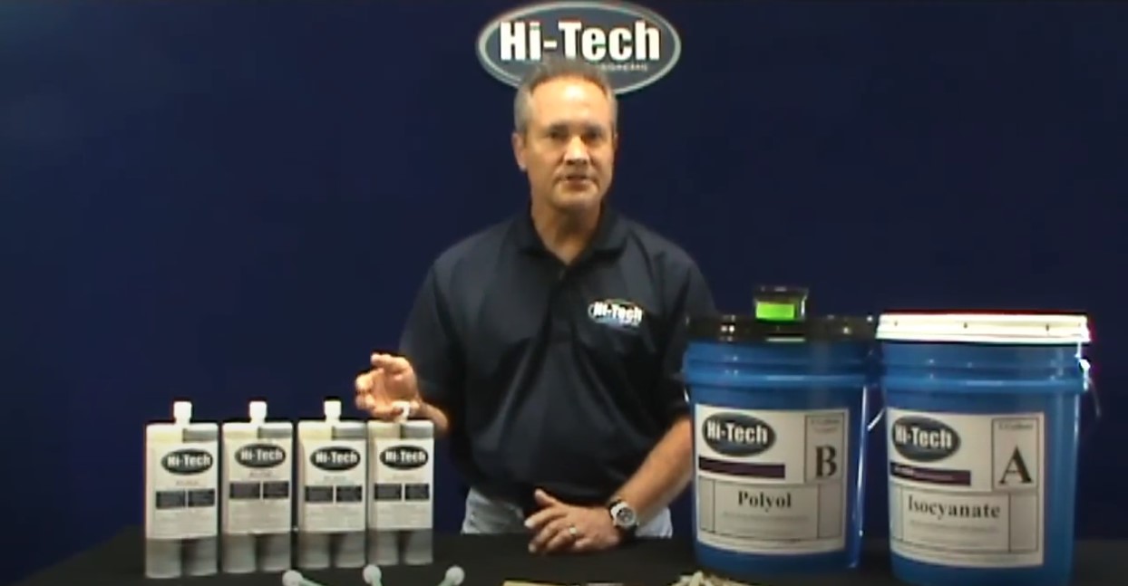 A man stands in front of a blue backdrop displaying the Hi-Tech logo, presenting two large containers labeled "Polyol" and "Isocyanate" on a table. Several smaller bottles and tools are arranged in front of him, likely for a demonstration.