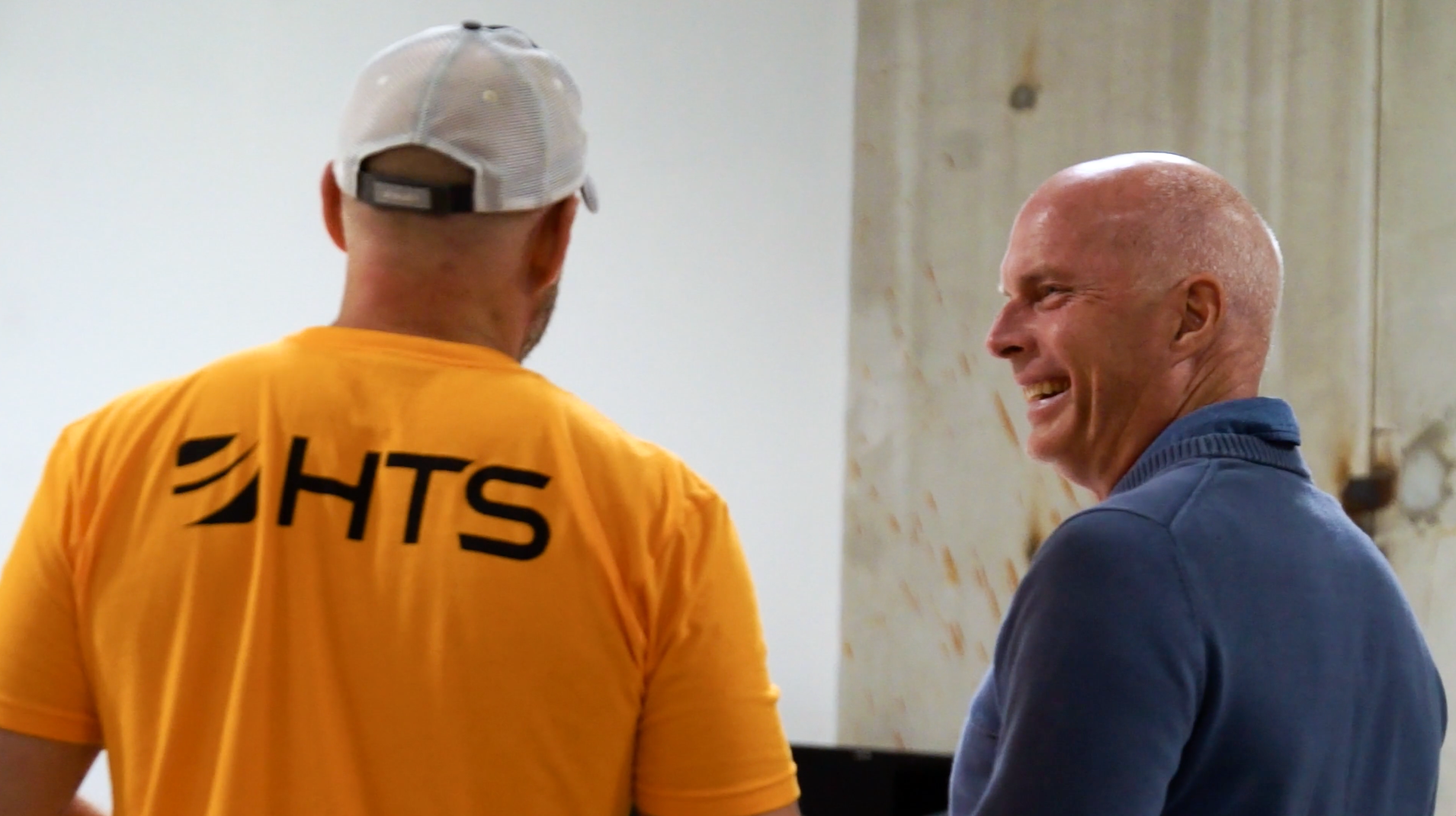 Two men are engaged in conversation, with one wearing a bright yellow shirt featuring the letters "HTS" on the back. The other man, smiling, is dressed in a blue sweater. They appear to be in a casual setting with a plain background.