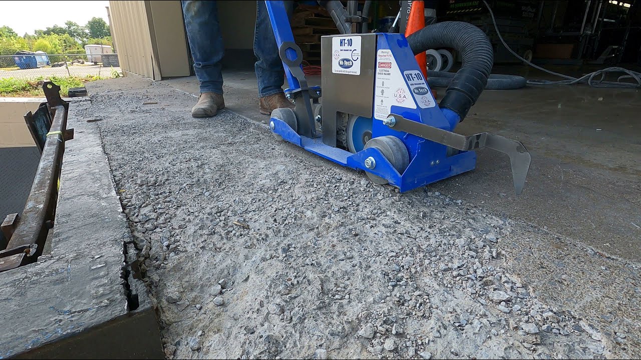 A blue concrete grinder is positioned on a partially finished concrete surface, with a person's legs visible nearby. The ground is rough and uneven, showing recent grinding work. Bright orange cones are in the background, indicating the work area.