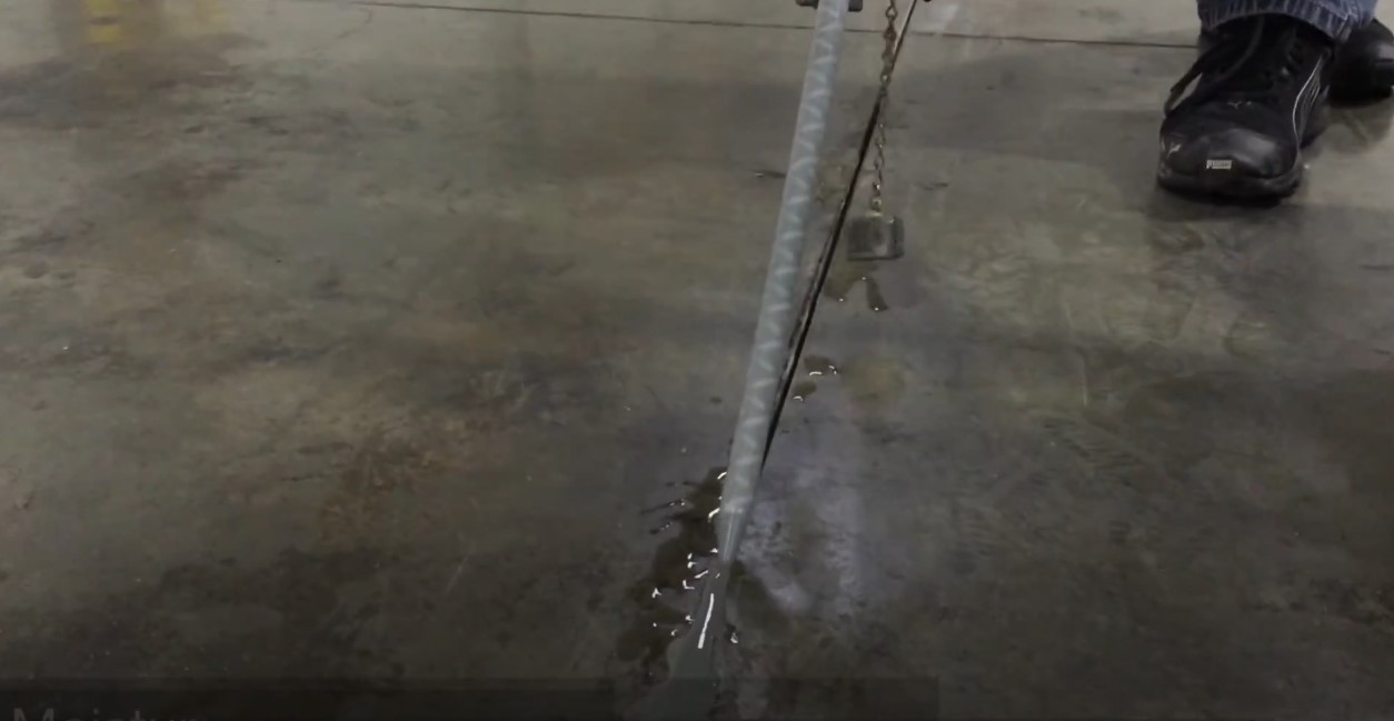 A metallic pole with a weight hangs above a puddle of water on a concrete floor. Water drips from the pole, creating ripples in the puddle. A black shoe is visible nearby.