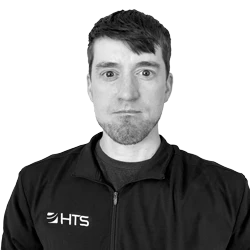 A man with short hair is wearing a black zip-up jacket with the letters "HTS" visible on the left side. He has a neutral expression and is facing the camera against a plain background.
