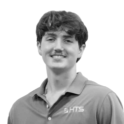 A young man with curly dark hair and a mustache smiles while wearing a red polo shirt with the logo "HTS." The background is plain and dark.