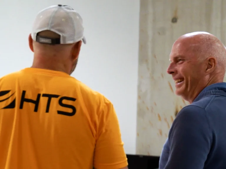 Two men are engaged in conversation, with one wearing a bright yellow shirt featuring the letters "HTS" on the back. The other man, smiling, is dressed in a blue sweater. They appear to be in a casual setting with a plain background.