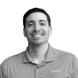 A smiling man with dark hair and a neatly trimmed beard, wearing a gray polo shirt with a logo on the left side. The background is plain black.